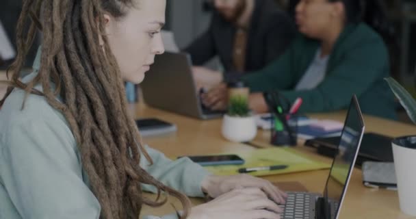 同僚がバックグラウンドで働いている間 ノートパソコンを使用してノートブックに書く若い女性の肖像画 現代の技術と職場の概念 — ストック動画