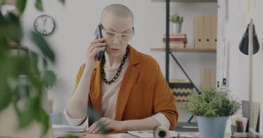 Neşeli işyeri sahibi cep telefonuyla konuşuyor. Gülüyor ve ofiste dizüstü bilgisayarla çalışıyor. Moda giysiler ve aksesuarlar giyen genç kadın işine odaklandı.