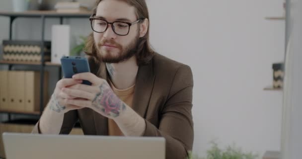 スマートフォンのテキストメッセージを使用し オフィスで屋内で笑顔で入れ墨の手を持つ陽気な若い男 コミュニケーションと現代技術の概念 — ストック動画