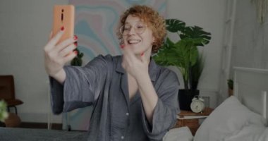 Pijamalı neşeli bir kadının evdeki yatakta akıllı telefon kullanarak online video görüşmesi yaptığı portre. İletişim ve modern yaşam tarzı kavramı.