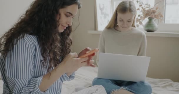 ベッドに座って電子機器と楽しい話をするガジェットのラップトップやスマートフォンを使用して幸せな友人 友情と近代的な技術の概念 — ストック動画