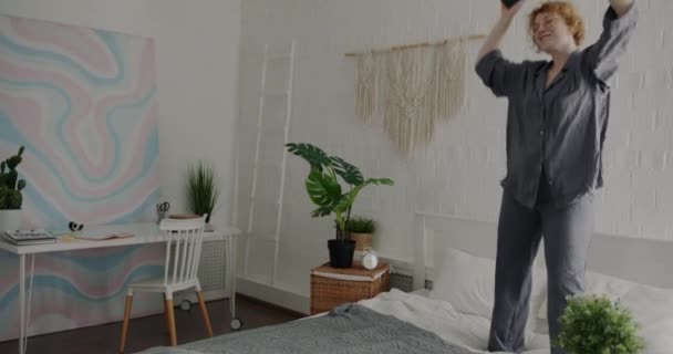 穿着睡衣在床上跳舞 拿着智能手机在家里玩乐的漂亮女人的慢动作肖像 人与现代生活方式概念 — 图库视频影像
