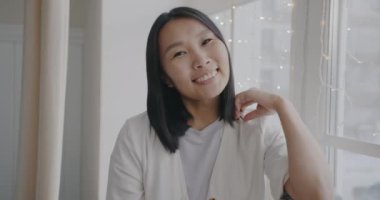 Güzel Asyalı bir kadının arka planda penceresi olan ve gülümseyen ve kameraya bakan yavaş çekim portresi. İnsanlar ve olumlu duygu kavramı.