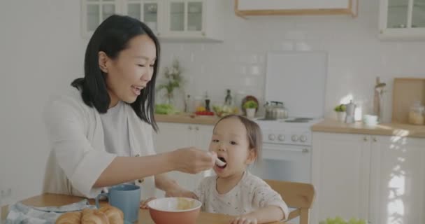 幸せなアジアの女性は 小さな娘にベビーフードを食べさせ アパートのキッチンで子供を見て笑っています 栄養と家庭生活の概念 — ストック動画