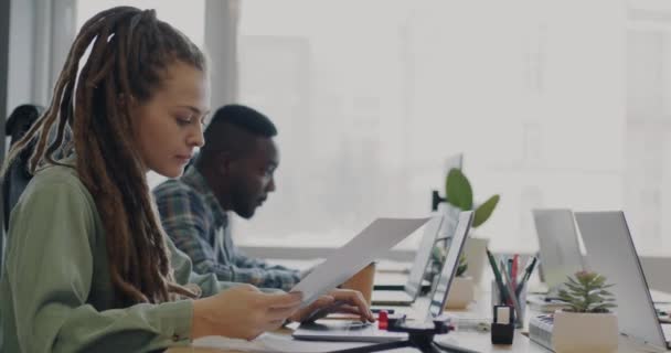 ラップトップで作業している若い女性は 男性の同僚にオフィスで仕事を議論する文書を与えます チームワークと成功したスタートアップコンセプト — ストック動画