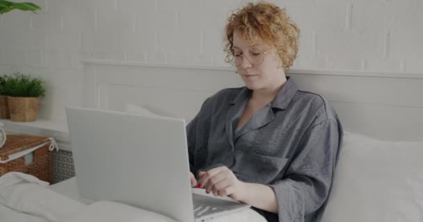 在家里穿着睡衣和笔记本电脑在床上工作的快乐的年轻女人的画像 上午使用计算机打字的女自由职业者 — 图库视频影像
