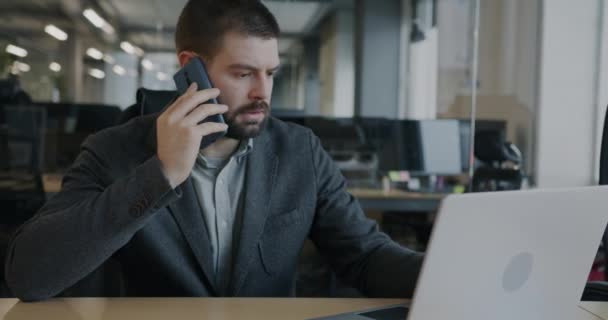 深刻なビジネスマンは 携帯電話で話をし ガラスの壁のオフィスで働くラップトップで遊んでいます ビジネスと近代的な技術の概念 — ストック動画
