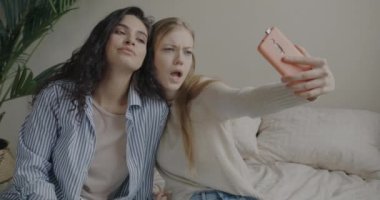 Neşeli genç bayan arkadaşlar akıllı telefon kamerasıyla selfie çekiyorlar evde oturmuş suratlarını buruşturarak eğleniyorlar. Teknoloji ve fotoğraf konsepti.