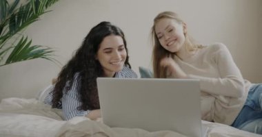 Neşeli kızlar online video görüşmesi yapmak için dizüstü bilgisayar kullanıyorlar. Yatakta uzanıp gülüyorlar ve arkadaşça sohbet ediyorlar. İnsanlar ve teknoloji kavramı.