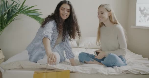 女性の友人に買い物袋に新しい服を示す幸せな若い女性が自宅でベッドに座っている 友情と現代のライフスタイルのコンセプト — ストック動画