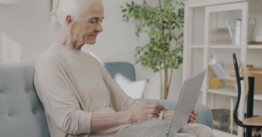 Üst düzey bir bayanın dizüstü bilgisayarla yazı yazıp evdeki kanepede oturan ekrana baktığı bir portre. Modern teknoloji ve emeklilik kavramı.