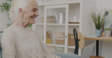 Evde dizüstü bilgisayarla konuşan ve gülümseyen yaşlı bir kadının portresi. Modern teknoloji ve iletişim konsepti.