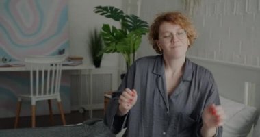 Evde müzik dinleyen, kablosuz kulaklık takan ve yatakta dans eden genç bayanın portresi. Modern teknoloji ve eğlence konsepti.