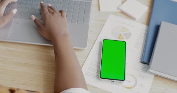 机の上にノートパソコンで入力する緑の画面のクロマキースマートフォンや女性の手のクローズアップ 現代のデバイスとモバイルアプリケーションの概念 — ストック動画
