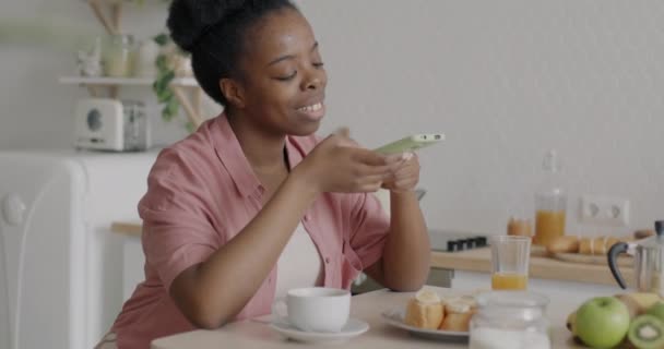 スマートフォンを使ったパジャマ姿の楽しそうな女の子と 台所のテーブルでガジェットを楽しみながら笑顔で座っています 電子機器やミレニアル世代の概念 — ストック動画