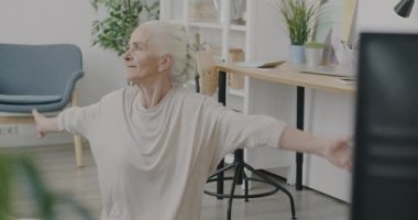 Oturma odasında spor yapan ve spor programı izleyen yaşlı bir kadın. Motive edilmiş yaşlı sporcu ve kitle iletişim araçları konsepti.