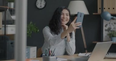 Ofis odasında eğlenen akıllı telefon kamerasıyla selfie çeken kaygısız Asyalı kadının yavaş çekim portresi. Neşeli milenyum ve fotoğraf konsepti.