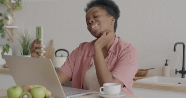 自宅のキッチンでノートパソコンや食品とテーブルに座ってスマートフォンで自撮りを取る喜びのビジネス女性 アフリカ系アメリカ人女性のポーズと笑顔が楽しい — ストック動画