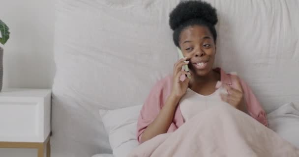 快乐的非洲裔美国女士在手机上微笑着坐在床上 享受着非正式的交流 人与智能手机概念 — 图库视频影像