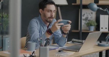 İşadamı, iş yerinde masasında oturmuş akıllı telefonuyla online video oyunu oynuyor. Teknoloji ve elektronik cihaz kavramı.