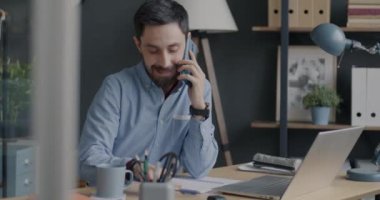 Ofisteki dizüstü bilgisayara bakarak cep telefonuyla konuşan ve not defterine bilgi yazan neşeli iş adamı. Halk ve profesyonel iletişim konsepti.