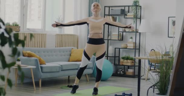 穿着时髦运动服独自在家锻炼的健康老妇人的画像 在客厅里 资深女运动员伸出双臂 站在瑜伽垫上 — 图库视频影像