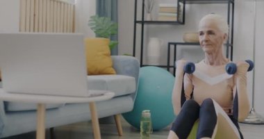 Spor kıyafetleriyle spor yapan yaşlı bir bayanın ağır çekimde portresi. Evde spor yapan dizüstü bilgisayara bakıyor. Sağlıklı yaşam tarzı ve emeklilik kavramı.
