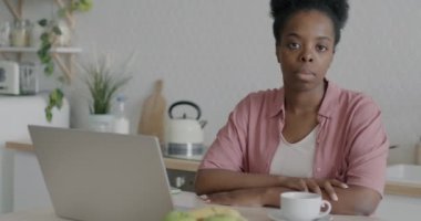 Ciddi bir Afro-Amerikan kadının portresi mutfak masasında dizüstü bilgisayarla oturmuş internetten çalışıyor ve kameraya bakıyor. Uzaktan eğitim ve zeki insanlar kavramı.