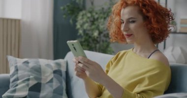 Akıllı telefon sörfü yapan neşeli genç bir bayan evdeki internet iletişiminin keyfini çıkarıyor. Modern cihazlar ve çağdaş yaşam tarzı kavramı.