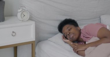 Yatakta uyuyan güzel Afro-Amerikan öğrencinin yavaş çekimde portresi. Sonra uyanıp evdeki çalar saati kapatıyor. Sabah ve insanlar konsepti.