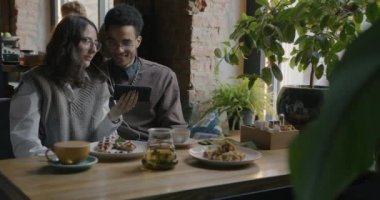 Akıllı telefondan komik videolar izleyen kız ve erkek öğrencilerin yavaş çekimde portresi kafedeki masada gülüyor. İnsanlar ve aygıtlar kavramı.