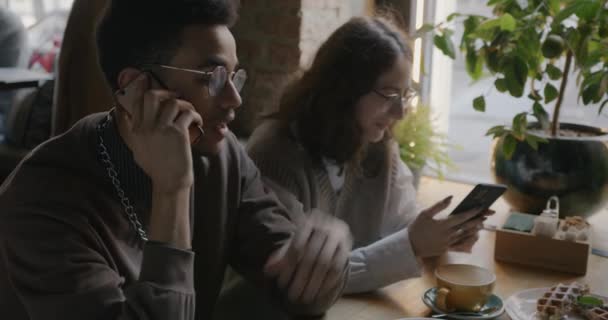 若い女性がカフェで食べ物とテーブルに座ってスマートフォンでテキストメッセージをしながら 楽しい男は携帯電話で話しています 通信とデバイスの概念 — ストック動画
