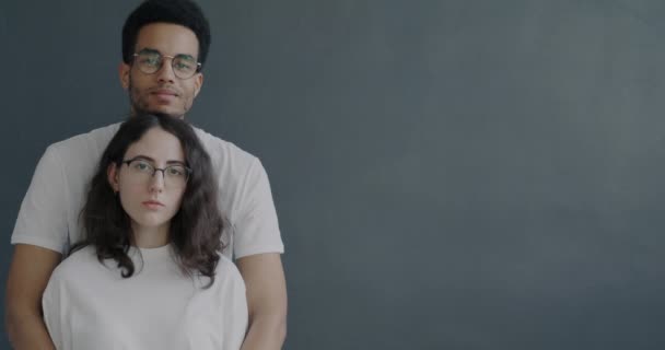 灰色の背景に立ってカメラを見て明るい笑顔で白人女性を抱きしめるアフリカ系アメリカ人男性の肖像画 人と感情の概念 — ストック動画