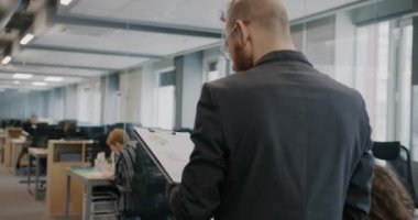 Takım elbiseli bir gencin modern açık uzay ofisinde doküman okuyup meslektaşlarını selamlarken çekilen Dolly fotoğrafı. İş adamları ve iş kavramı.