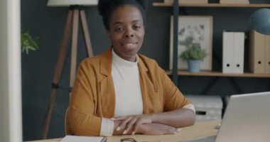 Ofiste oturmuş gülümseyen ve kameraya bakan başarılı Afrikalı Amerikalı kadının yavaş çekim portresi. İnsan ve iş kavramı.