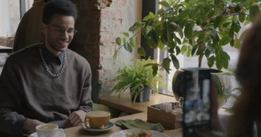 Kafe masasında kameraya poz veren mutlu Afro-Amerikalı adamın yavaş çekimleri. Bu arada kadın fotoğraf çekiyor. Halk ve fotoğraf konsepti.