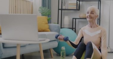 Modayı takip eden aktif giyimli yaşlı bir sporcunun portresi. Ev sağlığı için dizüstü bilgisayarda video izleyen dambıllarla spor yapıyor. Halk ve refah kavramı.