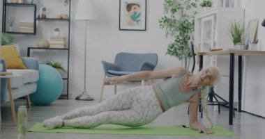 Evde yoga yaparak eğlenen aktif ve enerjik yaşlı bir kadının yavaş çekim portresi. Sağlıklı yaşam tarzı ve eğlence kavramı.