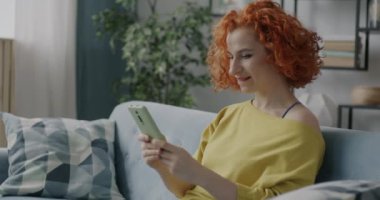 Akıllı telefondan güzel haberler alan heyecanlı genç bayan evdeki kanepede oturarak mutluluğu ifade ediyor. İnsanlar ve modern teknoloji kavramı.