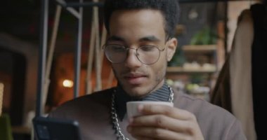 Genç Afro-Amerikan bir adamın portresi. Akıllı telefon kullanıyor ve kapalı bir kafede kahve içiyor. Modern teknoloji ve eğlence etkinliği kavramı.