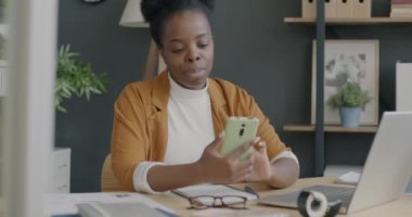 Ofiste çalışan neşeli Afro-Amerikan iş kadını akıllı telefon mesajları kullanıyor. Modern teknoloji ve profesyonel işgal kavramı.