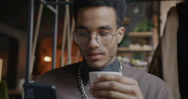 居心地の良いカフェでスマートフォンやコーヒーを屋内で使用して若いアフリカ系アメリカ人の男性の肖像画 現代技術とレジャー活動の概念 — ストック動画