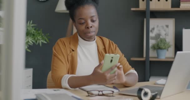 デスクでスマートフォンのテキストメッセージを使用して陽気なアフリカ系アメリカ人の実業家 現代の技術と職業概念 — ストック動画