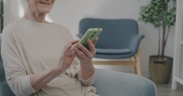 自宅で屋内でコンテンツを楽しむスマートフォンのタッチスクリーンを使用して陽気な高齢者の女性のスローモーションポートレート 古い時代と近代的な技術の概念 — ストック動画