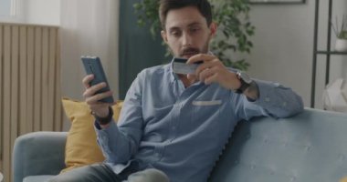 Akıllı telefon kullanarak online alışveriş yapan ve dairede kredi kartıyla ödeme yapan mutlu genç adam. Teknoloji ve e-ticaret kavramı.