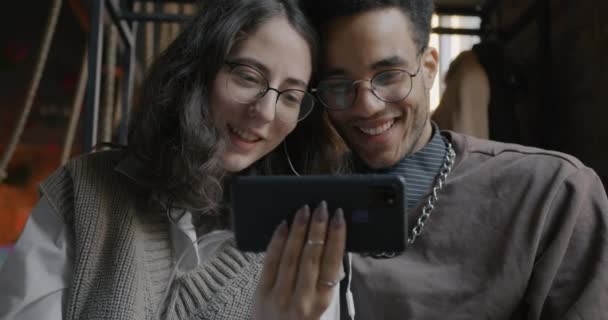 陽気な男性と女性は居心地の良いカフェでスマートフォンの画面上で面白いコンテンツを見て笑っています 二人で楽しそうに笑ってる — ストック動画