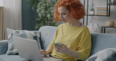 Dizüstü bilgisayar ve akıllı telefon kullanan genç bir kadının portresi. Dairedeki koltukta oturmuş iletişim kuruyor. Modern aygıt ve serbest çalışma konsepti.