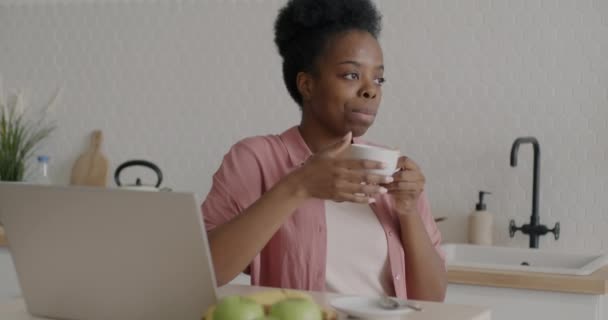 ノートパソコンとキッチンテーブルに座ってコーヒーを飲む若い女性の受動的なスローモーションの肖像画 千年と現代のライフスタイルのコンセプト — ストック動画