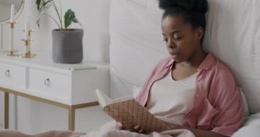 Güzel bir Afro-Amerikan kadının kitap okuyup evde edebiyattan zevk alırken gülümsemesinin yavaş çekimde portresi. Zeki insanlar ve yaşam tarzı konsepti.
