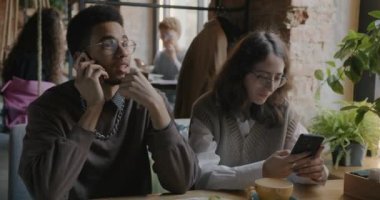 Kız arkadaşı kafede oturmuş akıllı telefon kullanırken cep telefonuyla konuşan genç adamın portresi. Araçlar ve iletişim konsepti.